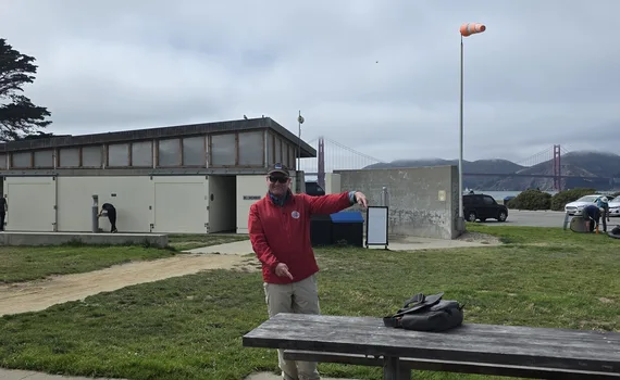 Jim, a San Francisco City Guides guide in a red jacket, pointing to the picnic tables by the changing rooms at Crissy Field East Beach, with the Golden Gate Bridge behind